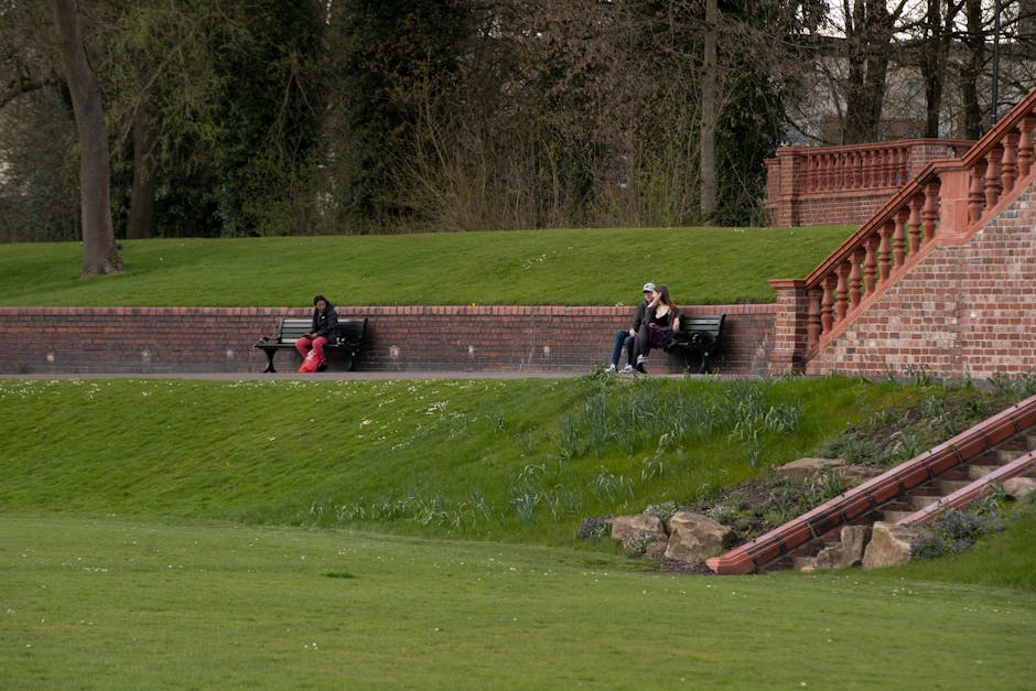 A landscaped sloped garden area at the Horniman Museum features well-maintained lush green grass and neatly trimmed borders. Two people are seated on dark green benches placed along a red brick retain