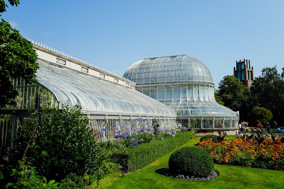 A landscaped sloped garden area at the Horniman Museum features well-maintained lush green grass and neatly trimmed borders. Two people are seated on dark green benches placed along a red brick retain
